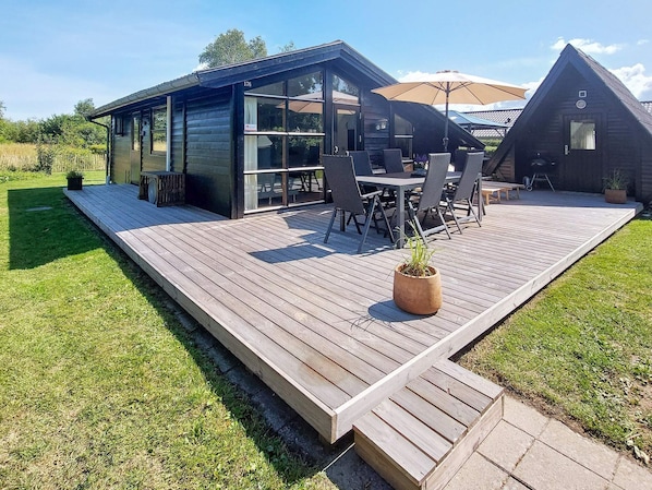 A modern outdoor deck with a dining area, a barbecue grill, and a small building with a glass door. - 6 Person Holiday Home in Hemmet (Hemmet)
