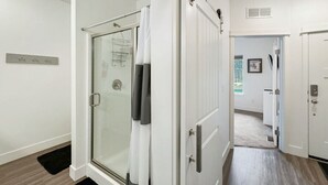 Une salle de bain moderne avec une douche à parois de verre, une porte blanche dotée d’un porte-serviettes et un couloir visible menant à une autre pièce.