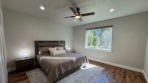 A bedroom with a bed, nightstands, a ceiling fan, a window, and a view of trees. - Stunning Home Close to Lake Cascade (Donnelly)