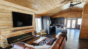 Een ruime woonkamer met een grote leren bank, een flatscreen televisie, een open haard en een keuken op de achtergrond.