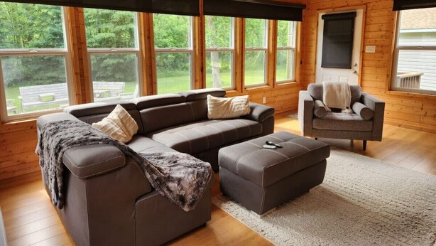 A cozy cabin interior with a sectional sofa, a loveseat, and a chair, all arranged around a large window offering a view of the outdoors. - The Birch Grove Escape (Gimli)