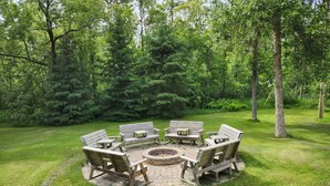 A circular outdoor seating area with wooden benches and a central fire pit, surrounded by a grassy lawn and trees. - The Birch Grove Escape (Gimli)