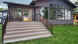 A modern house with a large glass door, a staircase leading to a deck, and a well-maintained lawn. - Boho Luxe Lakefront Cottage (Gimli)