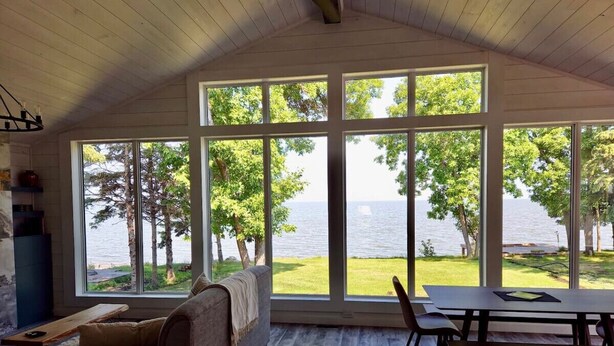 A room with large windows overlooking a body of water, a wooden ceiling, a sofa, a table, and a bench. - Boho Luxe Lakefront Cottage (Gimli)