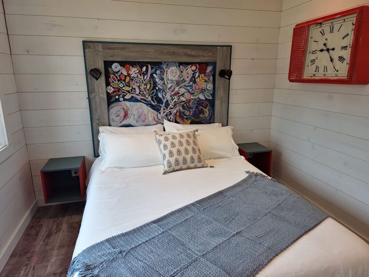 A bedroom with a bed, a headboard with a floral painting, a bedside table, a nightstand, and a wall clock.