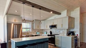 A modern kitchen with a central island, bar stools, stainless steel appliances, and a wooden ceiling. - Boho Luxe Lakefront Cottage (Gimli)