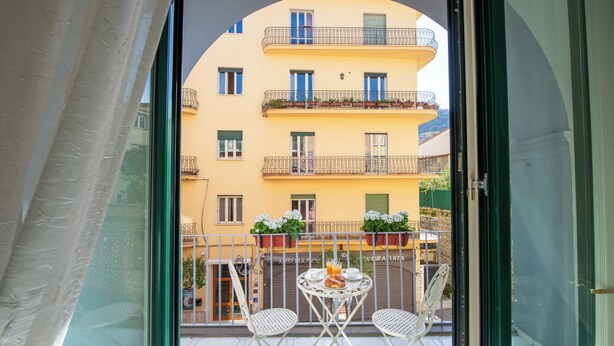 Ein Balkon mit Tisch und Stühlen, ein gelbes Gebäude mit Balkonen und ein Fenster mit Vorhang.