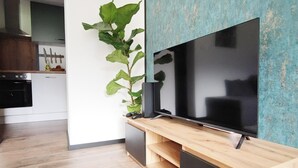 A modern living room with a flat-screen TV on a wooden stand, a large green plant, and a kitchen area in the background. - Great Apartment Near the ski Area (Schwendau)