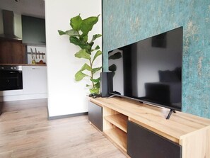 Un salon moderne avec un téléviseur à écran plat posé sur un meuble en bois, une grande plante verte et un coin cuisine à l’arrière-plan.