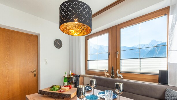A dining area with a wooden table set for two, a clock on the wall, and a window with a mountain view. - Great Apartment Near the ski Area (Schwendau)