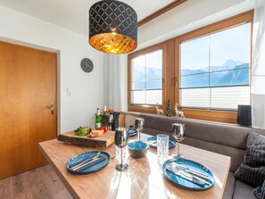 A dining area with a wooden table set for two, a clock on the wall, and a window with a mountain view. - Great Apartment Near the ski Area (Schwendau)