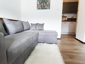 Un canapé d’angle gris avec des coussins, un tapis blanc à poils longs et une cuisine en arrière-plan.