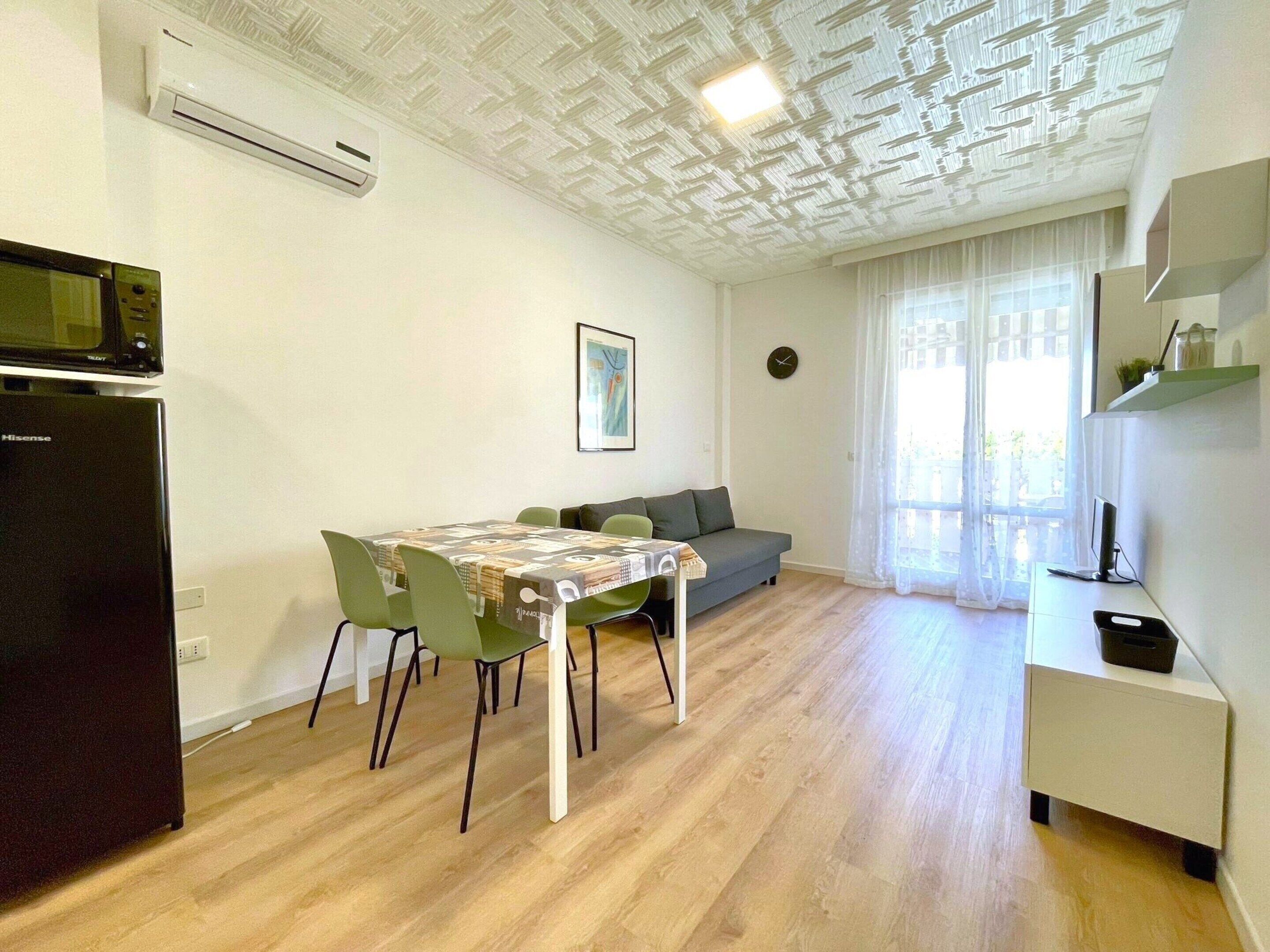 A modern apartment with a dining area, a kitchenette, a living space, and a large window with sheer curtains.