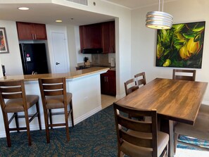A modern kitchen with dark wooden cabinets, a black refrigerator, and a central island. There is a dining area with a wooden table and chairs, and a large painting of flowers on the wall. - Hilton Hawaiian Village Lagoon Tower Oceanfront Premier 2-Bed, 2-Ba Condo (Honolulu)