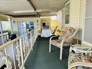 A covered balcony with a table and chairs, a trash bin, and a window with blinds. - Cozy Bright Park Model, Vibrant 55+ many Amenities (Mesa)