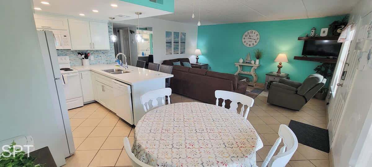 A modern kitchen with white cabinets, a dining area with a round table, and a living area with a brown sofa and a TV.