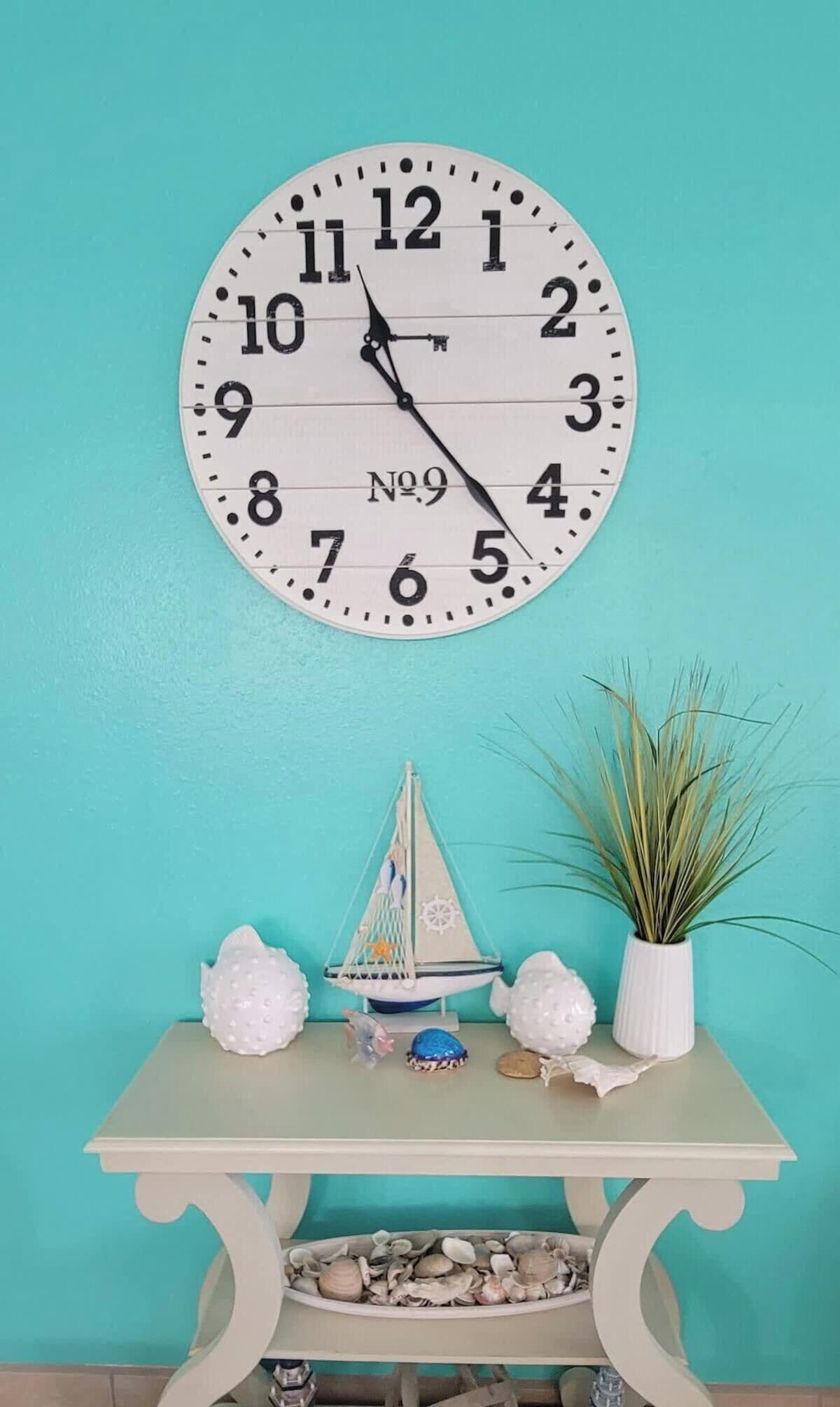 Eine Wanduhr mit maritimen Dekor, ein kleines Segelboot-Modell und ein Blumentopf auf einem Tisch vor einer türkisblauen Wand.