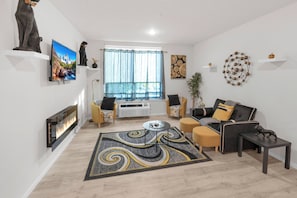 A modern living room with a fireplace, a large window with sheer curtains, a sofa set, a coffee table, and decorative items on the walls.
