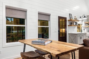A dining area with a wooden table and chairs, a book on the table, and a view of the outside through large windows with blinds.