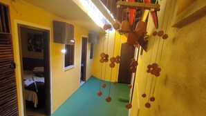 A room with a green carpet, yellow walls, and a wooden ceiling. There is a bed with a white blanket and a wooden headboard. A window with a curtain is visible, and a door leads to another room. - Pousada Caminho das Estrelas (Mata de São João)
