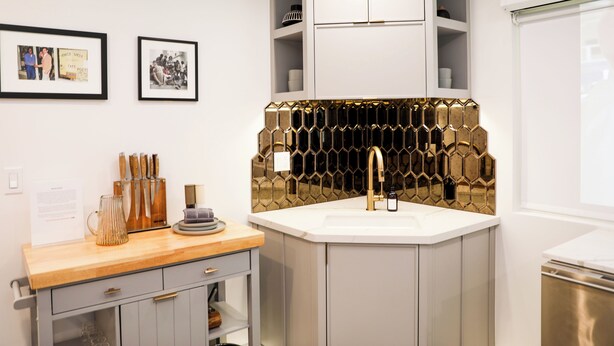 A modern kitchen with a white countertop, a gold-tiled backsplash, and a stainless steel sink. There are framed pictures on the wall and a wooden cabinet below the countertop. - The Escape - Suite 4 (Venice)