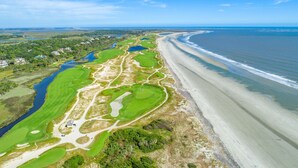 Vue aérienne d’un terrain de golf situé sur la côte, avec une plage et des zones résidentielles en arrière-plan.