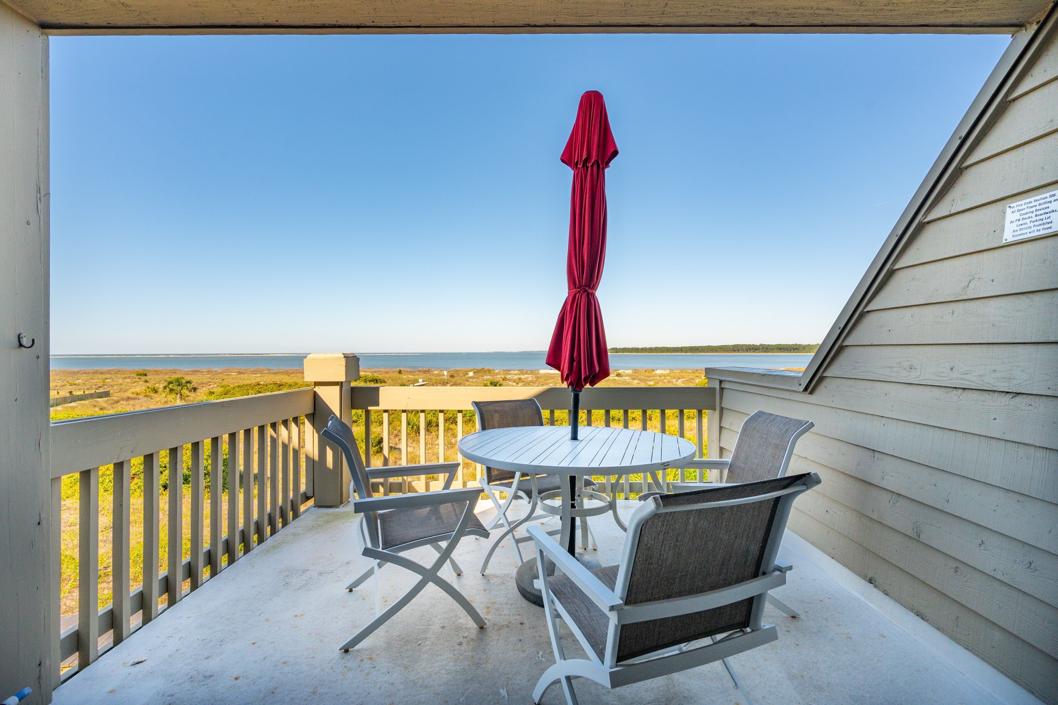 Un espace extérieur abrité, avec une table et des chaises, un parasol rouge et une vue sur la plage.