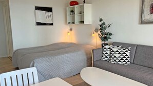 Moderni, hyvin valaistu huone, jossa on sänky, sohva ja pieni pöytä. Seinällä on hylly, jossa on koriste-esineitä, ja seinällä on myös kehystetty taulu.