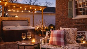 Un espace extérieur accueillant avec un banc, une table avec des verres et une bougie, et un jacuzzi installé sous une guirlande lumineuse.