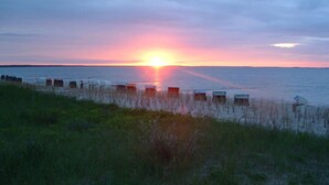 Une scène de plage avec un coucher de soleil sur l’eau, mettant en vedette plusieurs cabanes de plage et un premier plan herbeux.