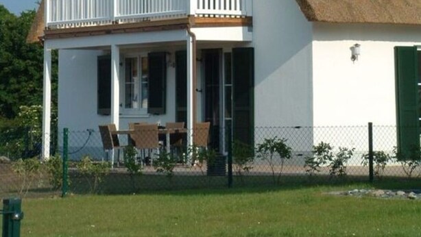 Une maison à deux étages avec un toit de chaume, un balcon doté de balustrades blanches et un petit espace extérieur aménagé avec des chaises.