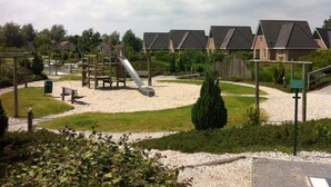 A playground with a slide and climbing structures, surrounded by a gravel area and greenery, with houses in the background. - Beachvilla Grachtenblik (Makkum)