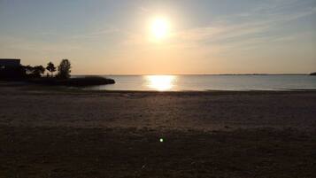 A beach with a sunset over the water, a sandy shore, and a distant shoreline with trees and a structure.