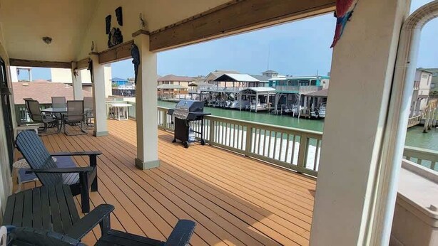A covered outdoor deck with wooden flooring, metal chairs, a grill, and a view of a marina with boats and buildings.