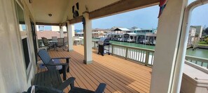 A covered outdoor deck with wooden flooring, metal chairs, a grill, and a view of a marina with boats and buildings.