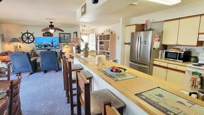 A compact kitchen with a central island, a dining area with blue chairs, and a living room with a TV and decorative items.