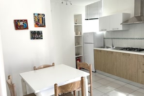 A kitchen with white cabinets, a white countertop, a stove, and a refrigerator. There are three chairs around a white table.