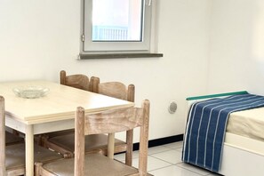 A room with a wooden dining table, four chairs, a small window, a bed with a striped blanket, and a white wall.
