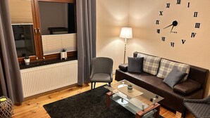 A living room with a sofa, armchairs, a glass coffee table, a lamp, a clock, and a window with curtains. - Schlossblick und Komfort in Stolpen (Stolpen)