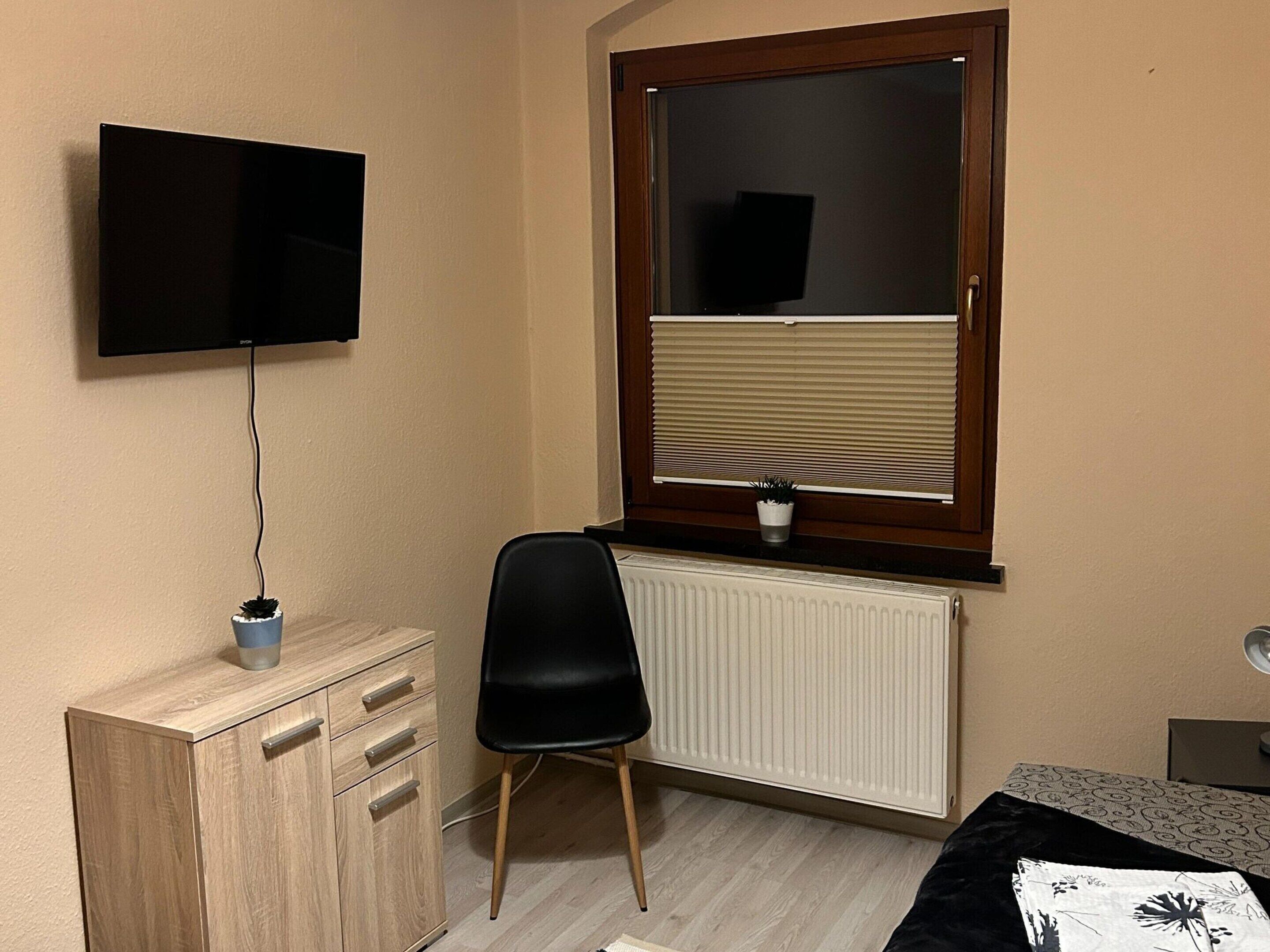 Une chambre avec une commode en bois, une chaise noire, un radiateur, une fenêtre avec des stores et un téléviseur fixé au mur.
