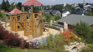 A wooden play structure with a red roof, surrounded by a stone wall and vegetation. - Cosy Apartment in Eibenstock (Eibenstock)