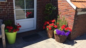 A brick house with a white door, two flower pots with red and purple flowers, and a small wooden bench to the left. - Tini - Haus Deichperle (Dornum)