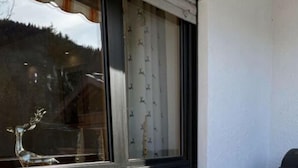 Un pequeño espacio al aire libre con mesa de madera y dos sillas blancas mullidas, una ventana con cortinas y una parrilla.