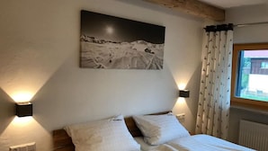 Un dormitorio con cama, dos lámparas de pared, un cuadro de un paisaje nevado y una ventana con cortinas.