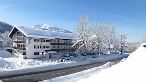 Un hotel de varios pisos con un paisaje nevado, una carretera al frente y árboles en el fondo.