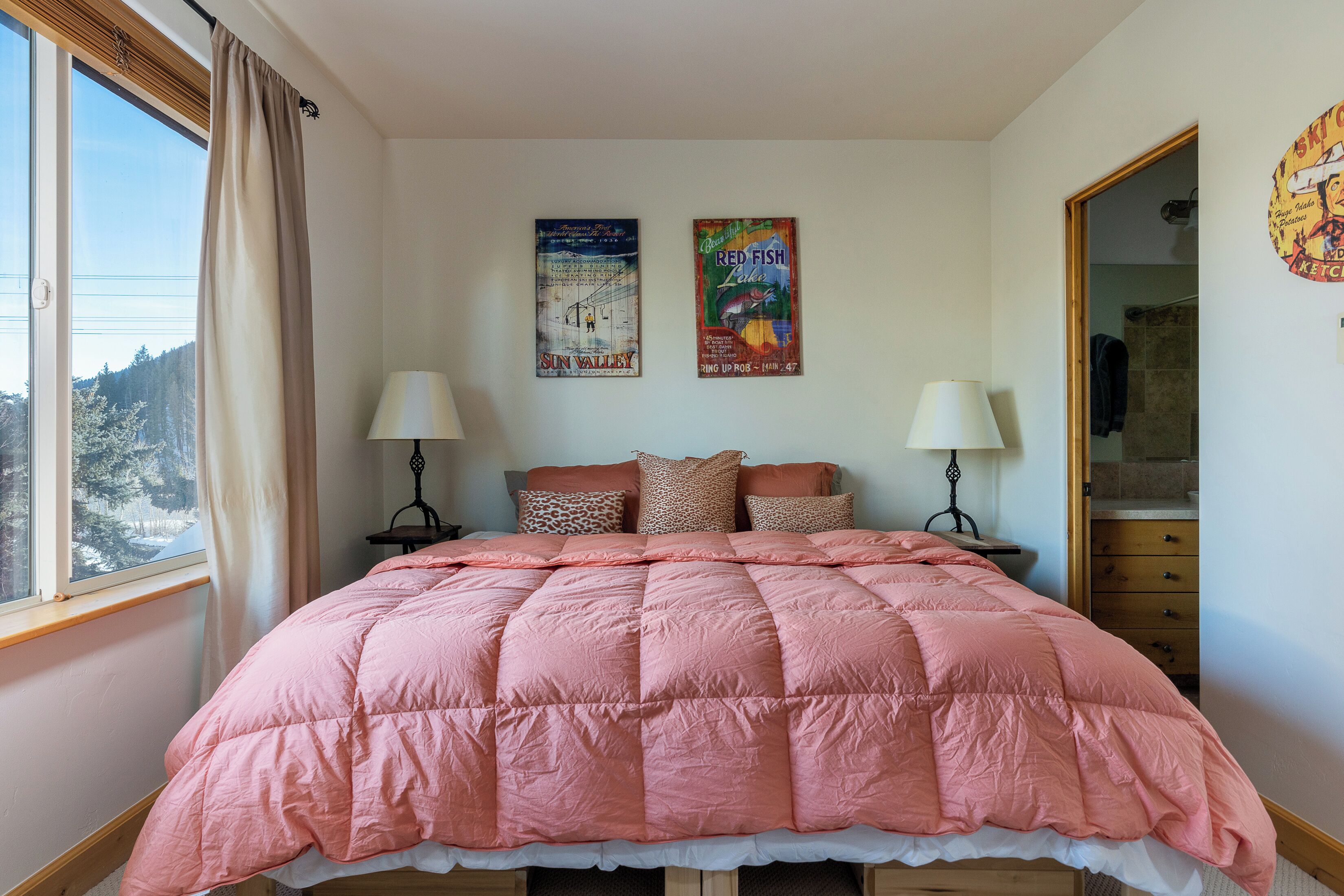 Une chambre à coucher avec une couvre-lit rose matelassé, deux lampes de chevet, une fenêtre avec des rideaux et deux œuvres d’art murales.