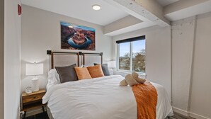 Una cama bien hecha con edredón blanco y manta naranja, una mesita de noche con lámpara y reloj, un cuadro sobre la cama y una ventana con vistas a árboles.