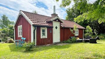 A red wooden house with a white door, a chimney, and a small porch with blue chairs.