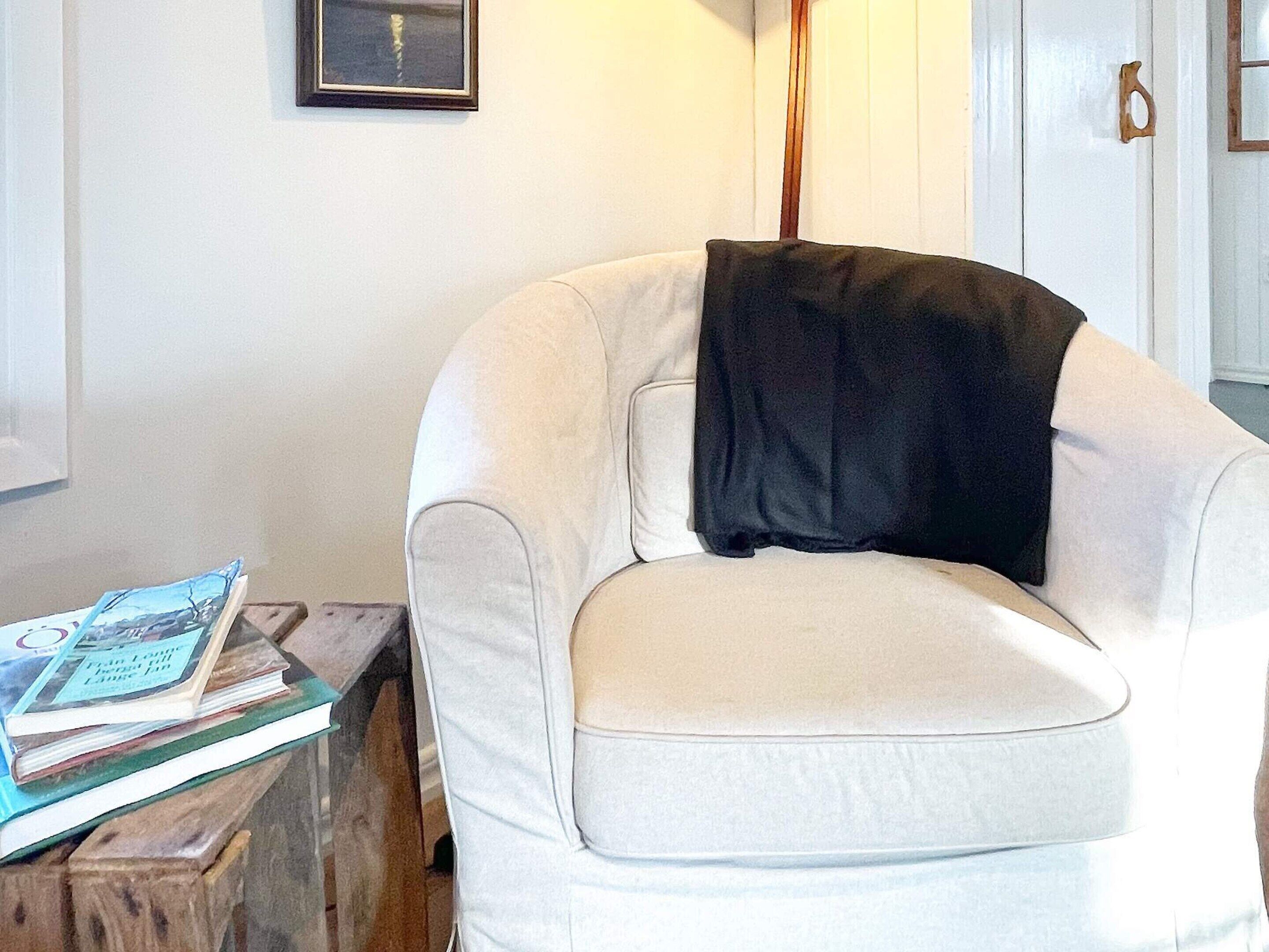 Un sillón blanco con una manta negra, una mesita de madera con libros y un cuadro en la pared.