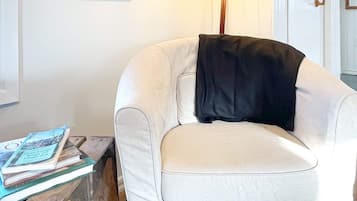 Un sillón blanco con una manta negra, una mesita de madera con libros y un cuadro en la pared.